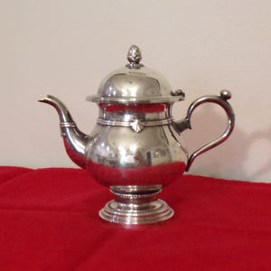 May include: A silver teapot with a rounded body, curved spout, and a loop handle. The lid is topped with a decorative finial. The teapot sits on a tiered base. The background is a solid red surface.