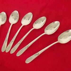 May include: Six silver spoons with oval bowls and long, slender handles. Each handle features a decorative floral design. The spoons are arranged on a solid red background, creating a classic and elegant aesthetic.
