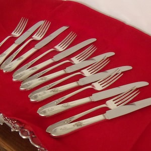 May include: A set of silver-colored forks and knives arranged on a red tablecloth. The silverware has decorative detailing on the handles. The image shows a formal dining setting.