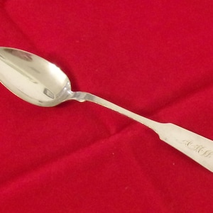May include: A polished silver-toned metal spoon with an oval bowl and a slender handle. The handle features an engraved monogram with the letters A.R.H. in a cursive script, ending in a rounded, slightly pointed tip with a subtle heart-like shape.