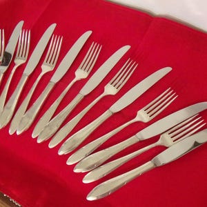 May include: A collection of stainless steel forks and knives arranged on a red surface. The silverware has a simple, elegant design, with smooth handles and a polished finish. The forks have four tines, and the knives have a curved blade.