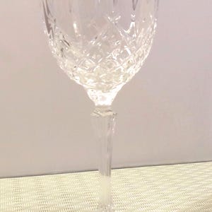 May include: A clear, crystal wine glass with a diamond-cut pattern on the bowl. The stem is slender and the base is round. The glass is empty and sits on a textured, light-colored surface.