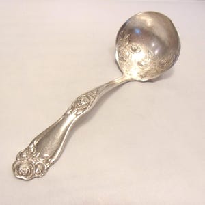 May include: A silver-colored ladle with a floral rose design on the bowl and handle. The handle curves gently, and the ladle appears to be made of metal. The ladle is likely for serving soups or sauces.