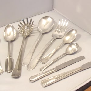 May include: A collection of eight silver-toned metal utensils including a ladle, a salad server, a large serving spoon, a dinner fork, three spoons of varying sizes, and two dinner knives with flat blades and polished handles.