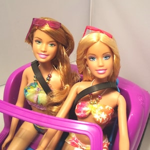 May include: Two blonde dolls wearing sunglasses and swimwear sit in a purple toy car. The dolls have fair skin, blue eyes, and are wearing colorful bikinis. One doll has pink sunglasses, and the other has red sunglasses. The car has a black seatbelt.