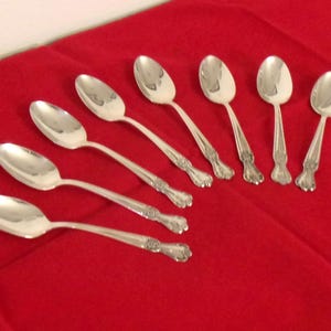 May include: A set of seven silver-toned metal spoons arranged in a diagonal pattern. Each spoon features a smooth, oval-shaped bowl and a slender handle that widens toward the base, displaying a subtle decorative embossed detail at the end.