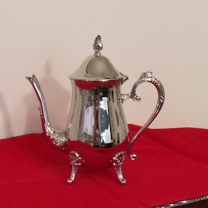 May include: A silver-colored tea or coffee pot with a curved spout and handle. The pot has a lid with a decorative finial and rests on three small feet. The pot is highly reflective, and sits on a red tablecloth.