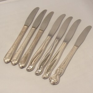 Puede incluir: Un juego de siete cuchillos de mantequilla plateados. Los cuchillos tienen una hoja curva y mangos ornamentados. Los mangos presentan dise&ntilde;os florales y de volutas. Los cuchillos est&aacute;n dispuestos diagonalmente sobre una superficie blanca.