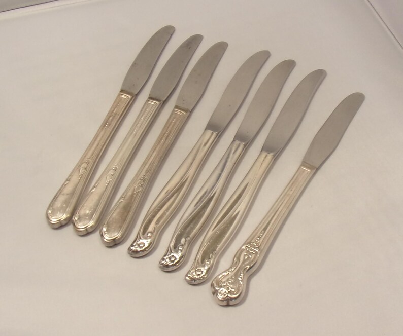 Puede incluir: Un juego de siete cuchillos de mantequilla plateados. Los cuchillos tienen una hoja curva y mangos ornamentados. Los mangos presentan dise&ntilde;os florales y de volutas. Los cuchillos est&aacute;n dispuestos diagonalmente sobre una superficie blanca.