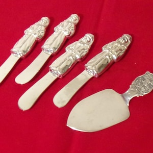 May include: A set of silver-colored serving utensils on a red background. The set includes four butter knives with a figure on the handle and a cake server with a decorative handle. The utensils are arranged on a red surface.