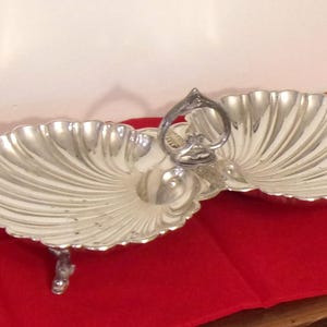 May include: A silver-toned metal serving dish featuring two conjoined shell-shaped bowls with a central heart-shaped handle. The piece is supported by small, ornate feet and has a polished, reflective surface with distinct radial ridges.