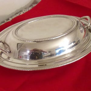 May include: A silver oval serving dish with a lid, featuring a rope-like border design. The dish has two small handles and sits on a red surface. The reflective surface of the dish shows a distorted reflection.