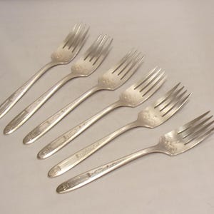 May include: Six silver forks arranged diagonally on a light gray surface. Each fork has four tines and a long, slender handle. The forks appear to be vintage, with some wear visible on the tines and handles.