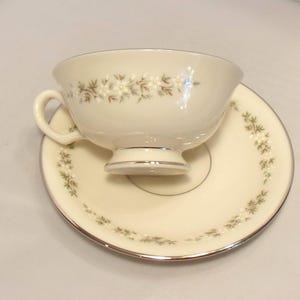 May include: Cream-colored teacup and saucer set with a floral design. The cup has a handle and sits on a matching saucer. Both pieces are trimmed with a silver-colored band. The floral pattern includes white and brown accents.