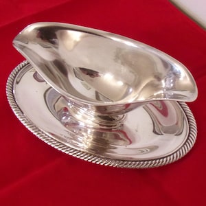May include: A silver gravy boat with an attached oval-shaped plate. The boat has a curved handle and a spout for pouring. The plate has a decorative rope-like border. The set is on a red surface.