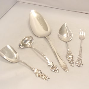 May include: A set of four silver-toned utensils including a wide serving spoon, a small ladle, a pointed spoon, and a three-tined fork. Each piece features a decorative floral pattern on the handle, with polished bowls and tines reflecting light.