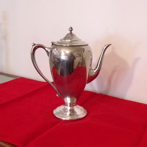 May include: A polished silver coffee pot with a lid and a curved handle. The pot sits on a pedestal base and has a long, slender spout. The pot is on a red tablecloth.