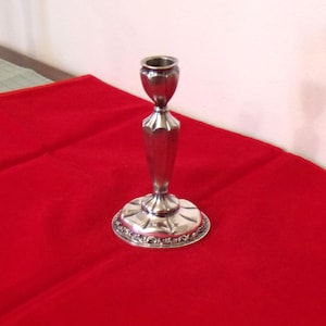 May include: A silver candlestick holder with a round base and a decorative floral pattern around the edge. The stem is faceted and tapers to a cup-shaped top. The candlestick is displayed on a red surface.