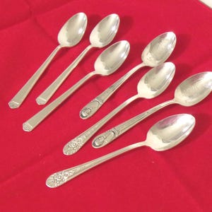 May include: Seven silver spoons arranged on a red surface. The spoons have oval bowls and long, slender handles. Some handles have a simple geometric design, while others feature ornate floral patterns. The spoons are reflective and appear to be made of metal.