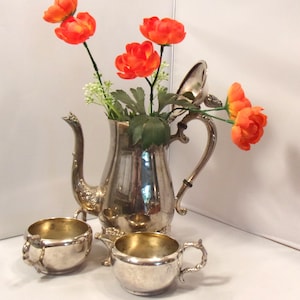 May include: A silver-plated tea set consisting of a tall teapot with an open lid and two matching bowls. The teapot is filled with bright orange artificial flowers with green leaves. The metal surfaces are reflective with ornate handle details.