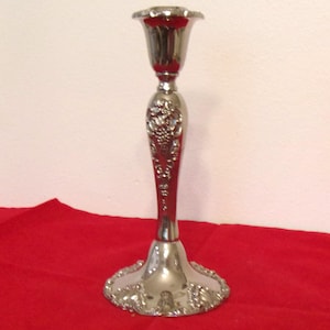 May include: A silver-toned metal candlestick holder featuring a flared top, a slender stem with embossed grape and vine detailing, and a wide, circular base with ornate, scalloped edges. The surface has a polished, reflective finish.