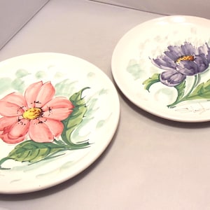 May include: Two round white ceramic plates featuring hand-painted floral designs. One plate displays a large pink flower with a yellow center and green leaves, while the other features a purple flower with a yellow center and green leaves.