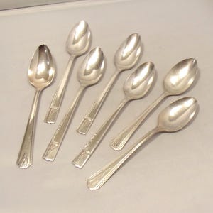 May include: A collection of seven silver spoons. The spoons have a classic design with oval bowls and long, slender handles. The handles feature decorative detailing near the bowl and at the end. The spoons are arranged on a light gray surface.