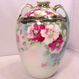 May include: A hand-painted ceramic vase with two handles, featuring a floral design of pink and white flowers with green leaves. The vase has a dark green band with gold accents near the top and gold detailing on the handles.