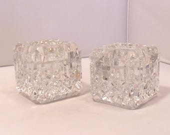 May include: Two clear, square crystal candle holders with a diamond-cut pattern. The candle holders are side-by-side, reflecting light and adding a touch of elegance. Ideal for home decor or special events.