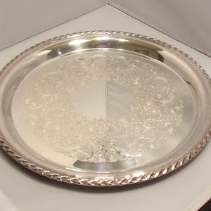 May include: A round, silver-colored serving tray with a decorative floral pattern in the center. The tray has a rope-like border. The tray is made of metal and reflects light.