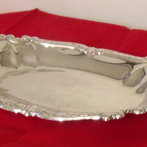 May include: A silver-colored, oblong serving tray with ornate, scalloped edges. The tray has a reflective surface and sits on a red fabric. The decorative tray is likely for serving food or displaying items.