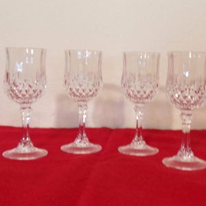 May include: Four clear crystal wine glasses with diamond-cut patterns. The glasses have a tulip-shaped bowl, a slender stem, and a round base. They are arranged on a red tablecloth against a neutral background.