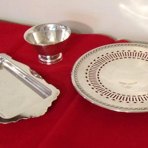 May include: A collection of silver serving pieces on a red surface. The set includes a rectangular tray with a scalloped edge, a small bowl, and a round plate with a decorative pierced design around the center.