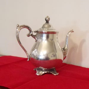 May include: A silver-colored teapot with a curved handle and spout, and a small lid with a decorative knob. The teapot sits on a decorative base and reflects the surrounding environment. The teapot is placed on a red tablecloth.