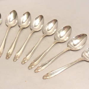 May include: A set of eight silver spoons arranged diagonally. Each spoon has a rounded bowl and a long handle with a decorative floral design near the bowl. The spoons are reflective, and the light catches their polished surfaces.