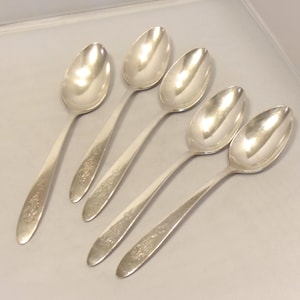May include: Five silver spoons with floral detailing on the handles. The spoons have a classic design with oval bowls and long, slender handles. The spoons are arranged on a light-colored surface, reflecting the light.