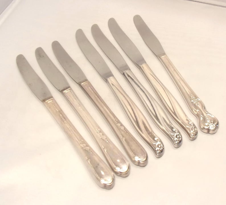 Puede incluir: Un juego de siete cuchillos plateados. Los cuchillos tienen una hoja lisa y brillante y un mango decorativo con dise&ntilde;os florales y de volutas. Los cuchillos est&aacute;n dispuestos en diagonal sobre una superficie blanca.