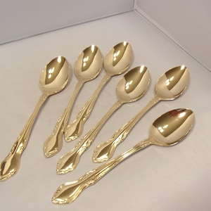 May include: Six gold-colored spoons with ornate handles are arranged on a light-colored surface. The spoons have a reflective, polished finish, and the handles feature a detailed floral design. The spoons are likely for serving or eating.