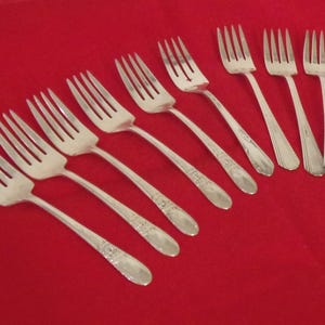May include: A collection of nine silver forks arranged diagonally on a red surface. The forks have tines and ornate handles. The image showcases a set of vintage silverware, ideal for formal dining or special occasions.