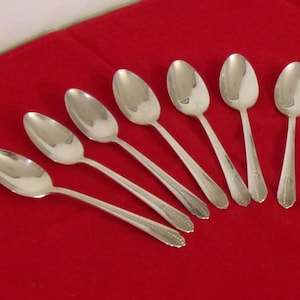 May include: Seven silver spoons arranged on a red surface. The spoons have oval bowls and decorative detailing on the handles. The spoons are likely for serving or eating.