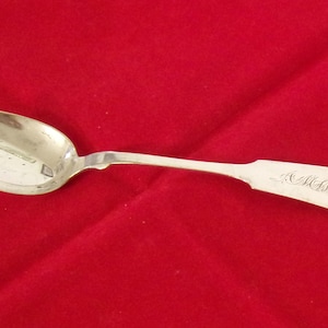 May include: A silver spoon with a rounded bowl and a slender handle. The handle has an ornate design and what appears to be engraved initials. The spoon is set against a red background.