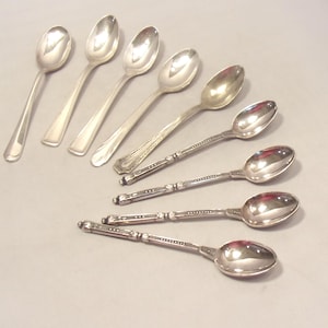 May include: A collection of eight silver-toned metal spoons arranged in a diagonal pattern. Four spoons feature plain, smooth handles, while the other four spoons have detailed, textured handles with small decorative bands and dotted patterns.