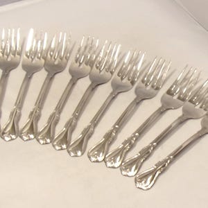 May include: A set of ten silver-toned metal forks arranged in a diagonal row. Each fork features four tines and a decorative, flared handle design with a smooth, polished finish. The forks are identical in shape and size, showing a consistent metallic sheen.