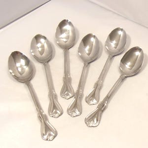 May include: A set of six polished silver-tone metal spoons arranged in a fan pattern. Each spoon features a smooth, oval-shaped bowl and a decorative handle with a flared, heart-shaped end and subtle embossed detailing near the base of the stem.