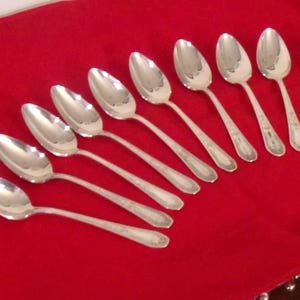 May include: Nine silver spoons arranged diagonally on a red tablecloth. The spoons have a classic design with oval bowls and ornate detailing on the handles. The spoons are reflective, and the red cloth provides a contrasting backdrop.