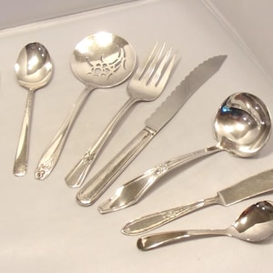 May include: An assortment of silver-colored flatware, including spoons, a fork, a knife, and a ladle. The utensils are arranged on a light gray surface, showcasing their reflective qualities. Some pieces feature decorative floral patterns.