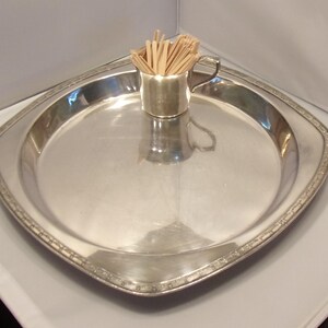 May include: A polished silver-toned metal serving tray with a square shape and rounded corners, featuring a decorative embossed border. A small silver-toned metal cup with a handle sits in the center, filled with a bundle of light-colored wooden toothpicks.