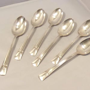 May include: Six silver spoons with oval bowls and decorative handles are arranged on a light gray surface. The spoons have a reflective, polished finish, suggesting they are made of stainless steel or a similar material. The handles feature a detailed design.