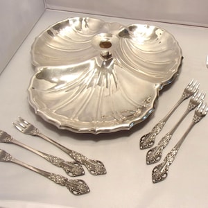 May include: A silver-toned metal serving dish shaped like a large scallop shell with a central handle, accompanied by five small forks featuring ornate floral embossed handles and one plain, slender silver-toned metal fork with a smooth handle.