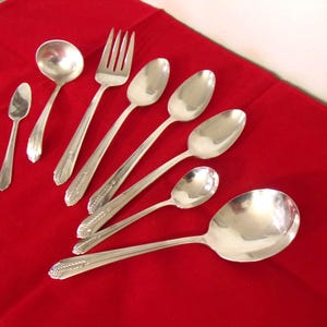 May include: A collection of silver-colored flatware, including spoons, a fork, and a ladle, arranged on a red surface. The utensils have decorative detailing on the handles. The arrangement includes various sizes of spoons.